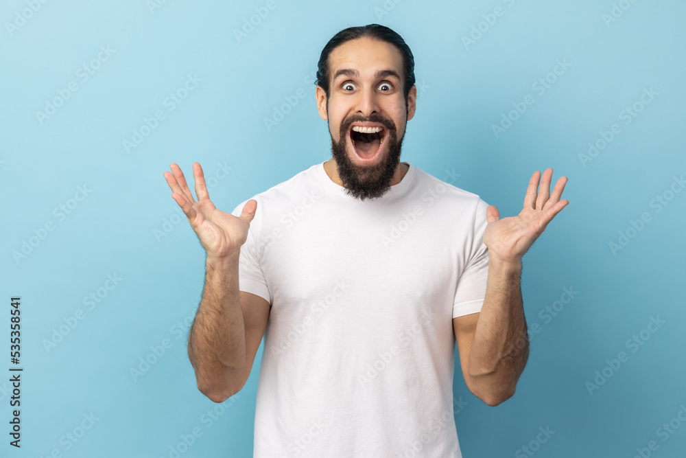 Portrait of man with beard wearing white T-shirt standing with mouth open in surprise, has shocked expression, keeping hands raised. Indoor studio shot isolated on blue background. Portrait of man with beard wearing white T-shirt standing with mouth open in surprise, has shocked expression, keeping hands raised. Indoor studio shot isolated on blue background.