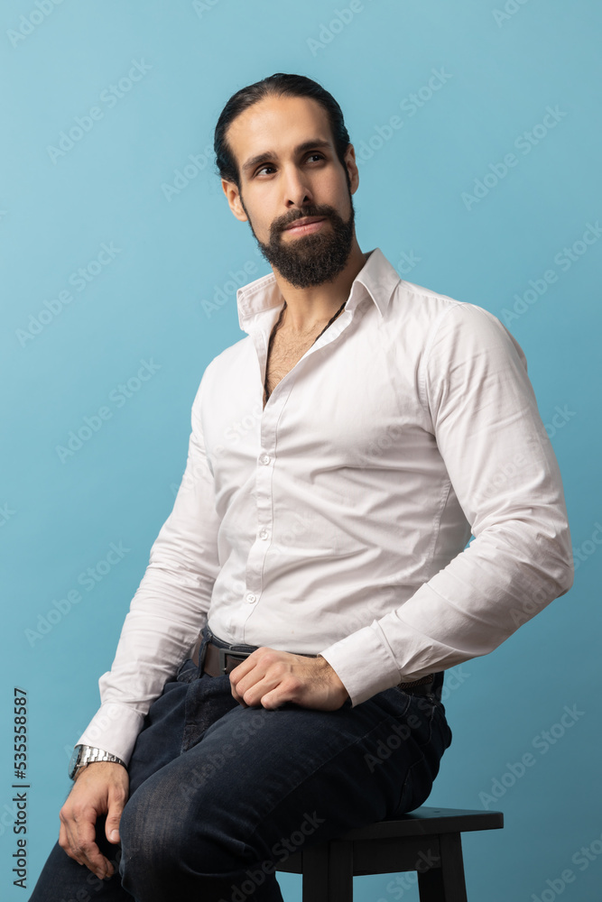 Obraz premium Unsmiling serious dark haired attractive man with beard wearing white shirt and black trousers, looking away with attentive look while sitting on chair. Indoor studio shot isolated on blue background.