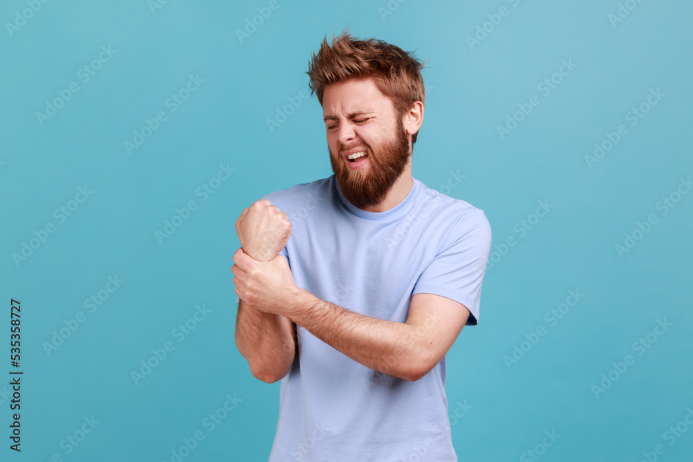 Portrait of unhealthy sick young adult bearded man massaging wrist feeling pain, muscle sprain, joint inflammation, tunnel syndrome. Indoor studio shot isolated on blue background. Portrait of unhealthy sick young adult bearded man massaging wrist feeling pain, muscle sprain, joint inflammation, tunnel syndrome. Indoor studio shot isolated on blue background.
