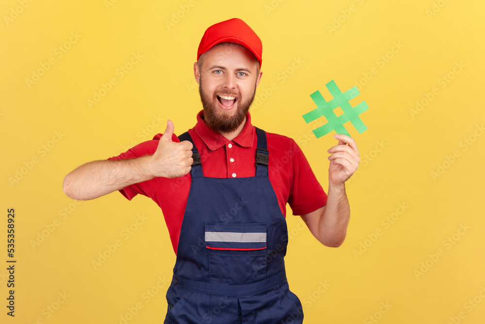 Portrait of excited handyman wearing blue overalls standing holding green hashtag and showing thumb up, looking at camera with smile. Indoor studio shot isolated on yellow background.