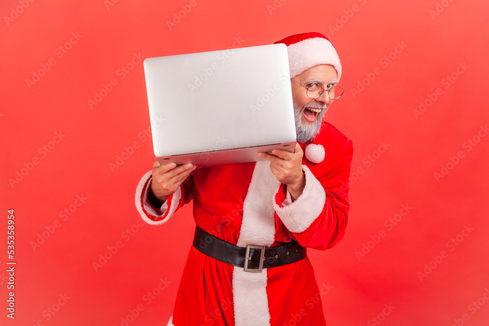 Φωτογραφία Portrait of positive elderly man with gray beard wearing santa claus costume posing peeking from laptop computer, laughing happily, winter holidays