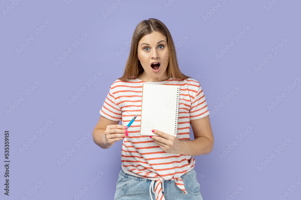 Portrait of amazed surprised blond woman wearing striped T-shirt holding organizer with empty paper with copy space for advertisement. Indoor studio shot isolated on purple background. Portrait of amazed surprised blond woman wearing striped T-shirt holding organizer with empty paper with copy space for advertisement. Indoor studio shot isolated on purple background.
