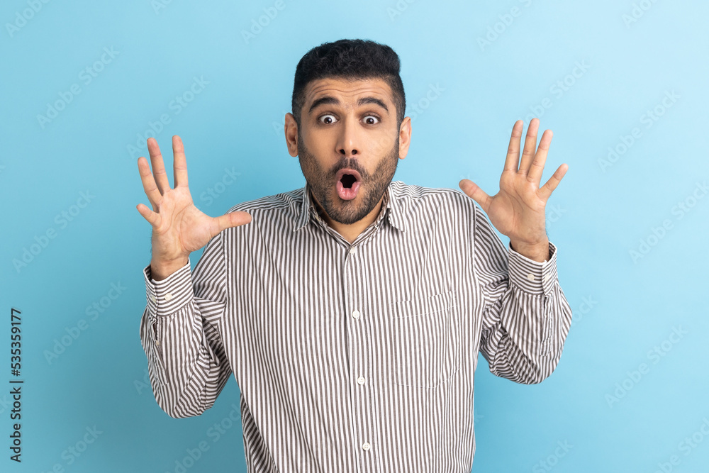 Fototapeta premium Portrait of surprised astonished bearded businessman standing with open mouth and raised arms, being impressed of shocking news, wearing striped shirt. Indoor studio shot isolated on blue background.