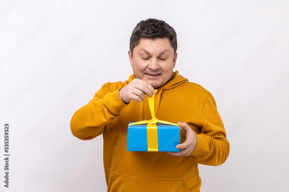Middle aged man opening gift box, unwrapping birthday present for celebrating holiday, pulling ribbon, looking at gift, wearing urban style hoodie. Indoor studio shot isolated on white background. Middle aged man opening gift box, unwrapping birthday present for celebrating holiday, pulling ribbon, looking at gift, wearing urban style hoodie. Indoor studio shot isolated on white background.