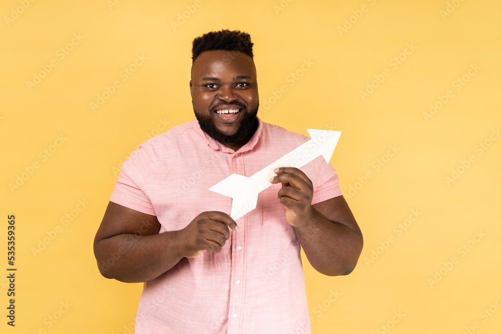 Portrait of smiling satisfied man wearing pink shirt pointing direction with arrow aside, looking at camera with positive expression. Indoor studio shot isolated on yellow background. Portrait of smiling satisfied man wearing pink shirt pointing direction with arrow aside, looking at camera with positive expression. Indoor studio shot isolated on yellow background.