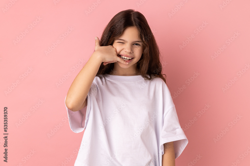 Portrait of smiling happy positive little girl wearing white T-shirt flirting holding fingers near ear showing call gesture, answering call. Indoor studio shot isolated on pink background. Portrait of smiling happy positive little girl wearing white T-shirt flirting holding fingers near ear showing call gesture, answering call. Indoor studio shot isolated on pink background.