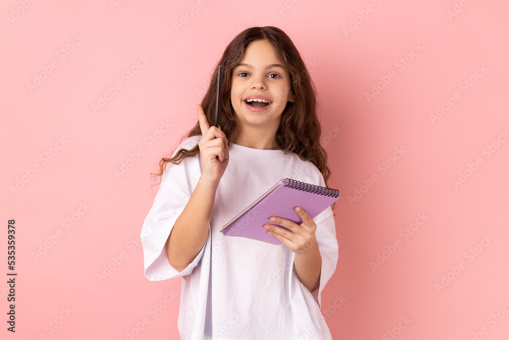 Portrait of excited charming little girl wearing white T-shirt writing in paper notebook and raising finger up, having new good idea. Indoor studio shot isolated on pink background. Portrait of excited charming little girl wearing white T-shirt writing in paper notebook and raising finger up, having new good idea. Indoor studio shot isolated on pink background.