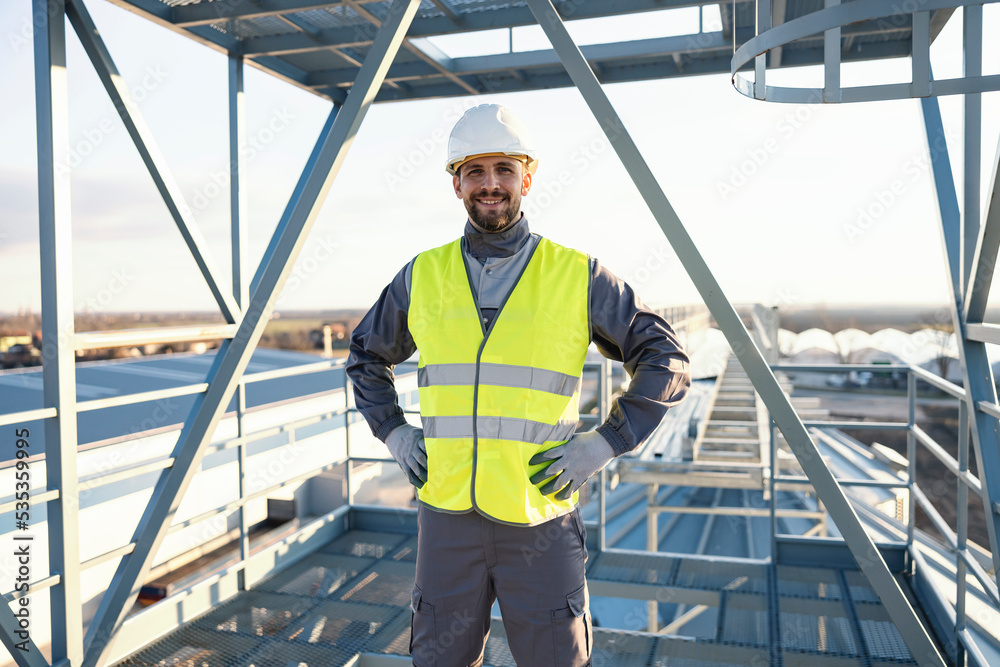 An industry worker stands on the metal construction at a height and smiles at the camera.