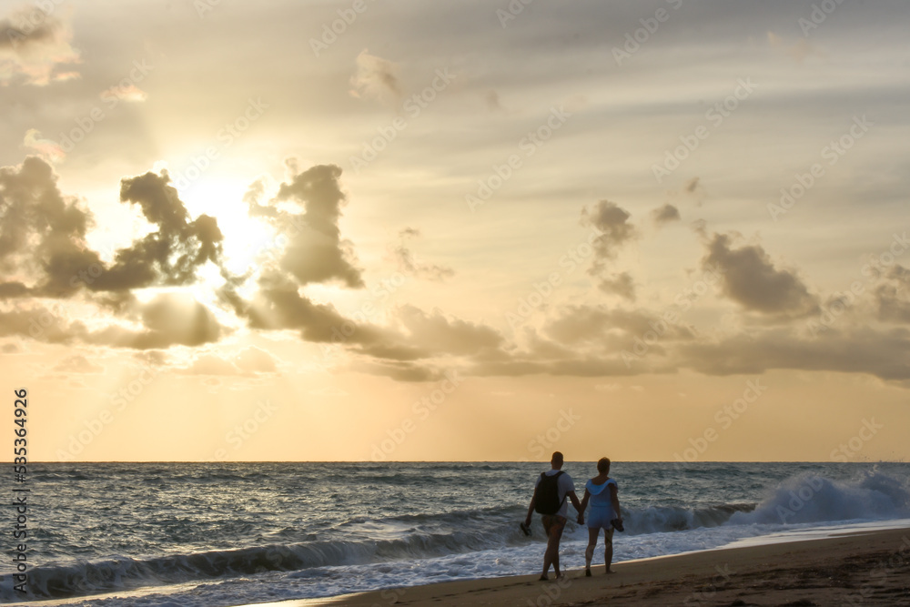 Young couple hand to hand walking on a beach at cloudy sunset