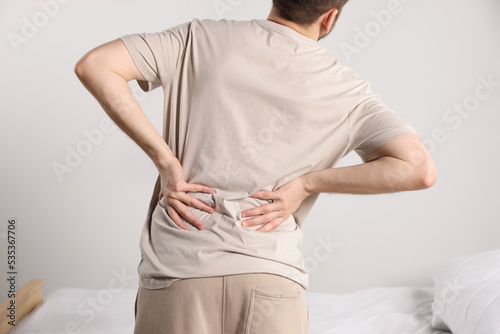 Man suffering from back pain while standing near bed in room, closeup. Symptom of scoliosis