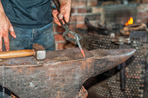 A forger wearing blue jeans and a t-shirt holds fire thongs and a metal twisted bolt on a vintage anvil. There's rounding hammer tool with a wooden handle in the blacksmith foundry shop with a furnace