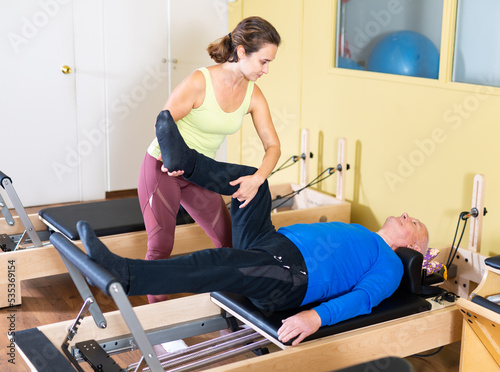 Focused positive senior man practicing pilates system on reformer to improve and maintain mobility under supervision of qualified female trainer