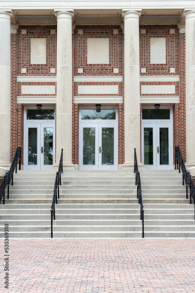 Concrete steps lead to a red double door of a historic building. The wall of the building is made of a light grey granite block. There are four metal handrails dividing the stairs to the entrance.