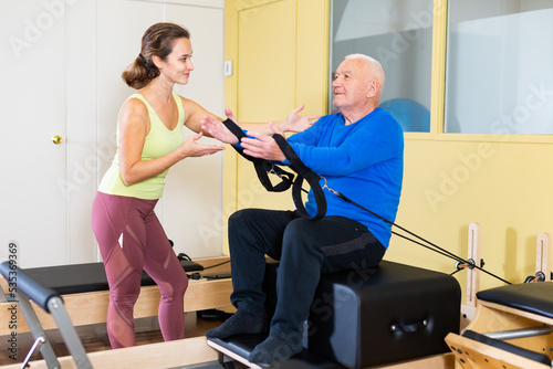 Personal female trainer controlling movements of senior man doing pilates on reformer in fitness studio. Healthy active lifestyle
