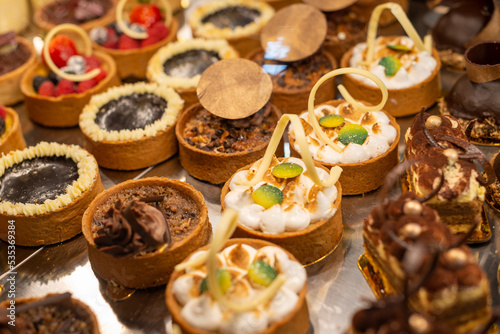 Rows of sweet pastries in a glass showcase at a patisserie. There are small round baked cakes, comfit, desserts, chocolate mousse, pies, biscuits, berry tarts, and fruit snacks on display for sale.