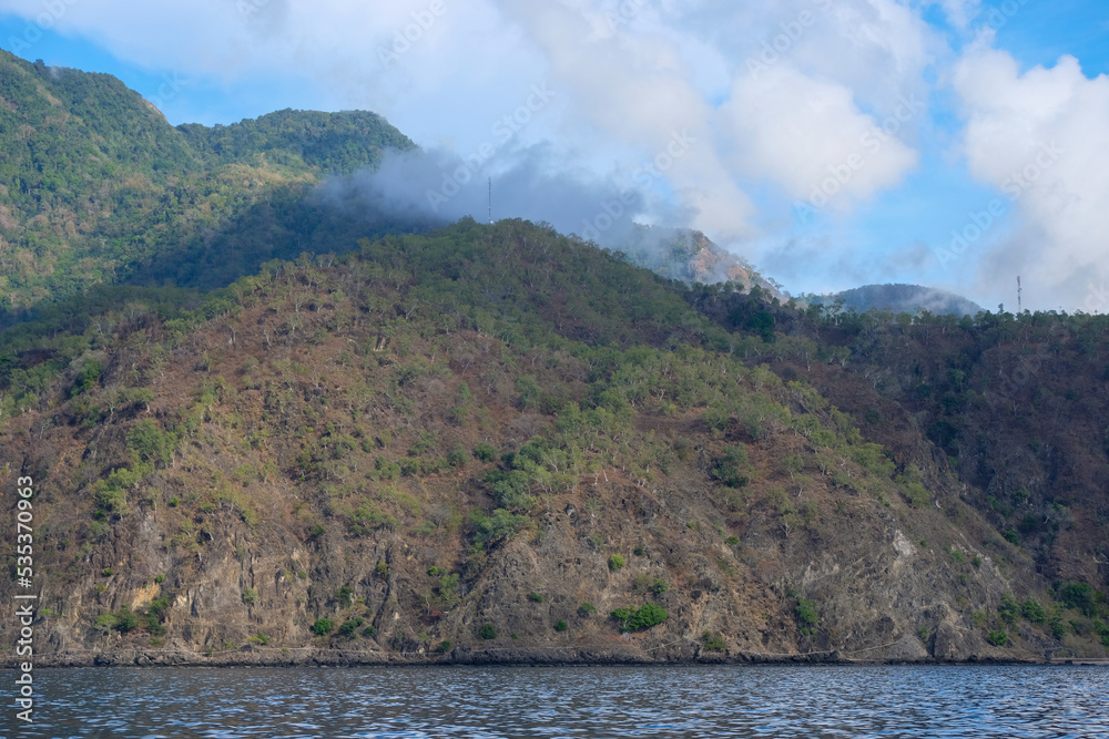 Rugged, steep sloped mountains, with sparse trees during the dry season, rising up from the ocean, and clouds lingering on mountaintops, on remote tropical Atauro Island in South East Asia Rugged, steep sloped mountains, with sparse trees during the dry season, rising up from the ocean, and clouds lingering on mountaintops, on remote tropical Atauro Island in South East Asia