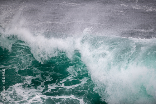 An angry turquoise green color massive rip curl of a wave as it rolls along the ocean. The white mist and froth from the wave are foamy and fluffy. The Atlantic Ocean in the background is deep blue