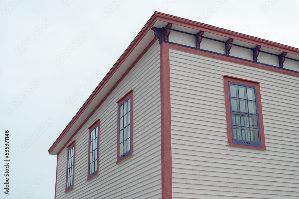 The roof section of a large white vintage wooden house with decorative pink and purple wood trim. There are three multi-pane windows on the top floor. The background is a dramatic blue cloudy sky.