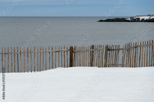 An old wooden twig fence with a small gate on the edge of a cliff. There's a view of the sea in the background and a meadow in front is covered in fresh white snow. The sky is blue and cloudy. 
