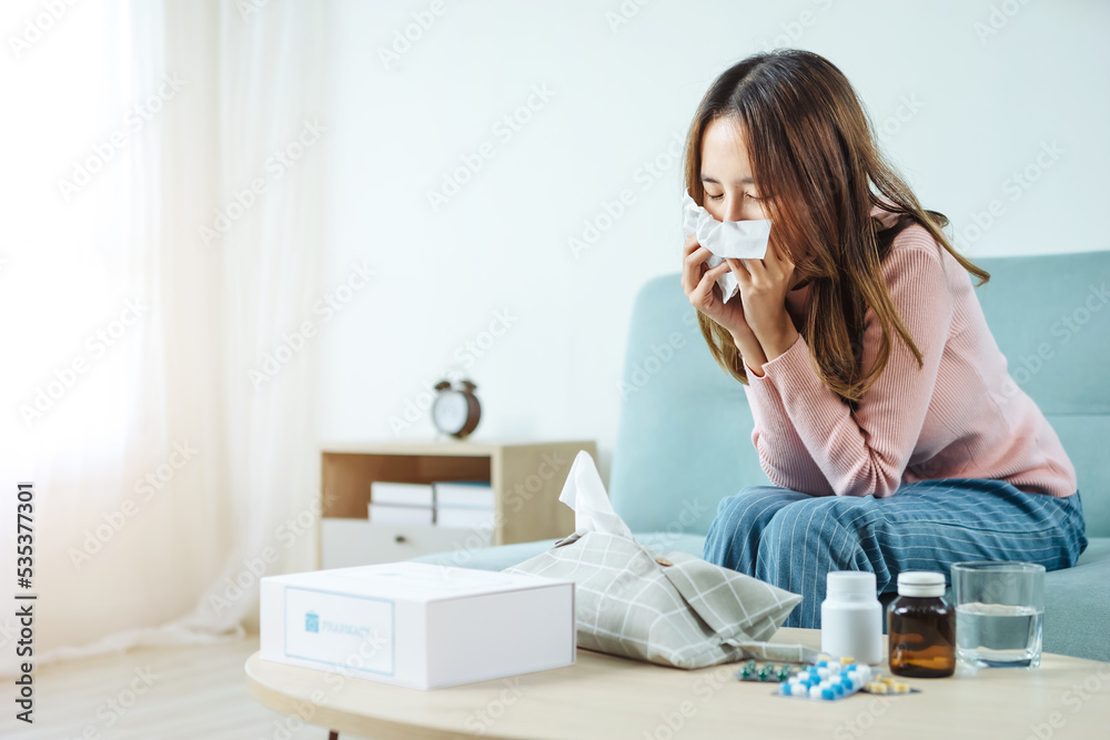 Young Asian woman sick, tired, she sitting and resting on blue fabric sofa and blanket after takes medicines pills in living room at her home. Woman use the phamacy first aid kit box Young Asian woman sick, tired, she sitting and resting on blue fabric sofa and blanket after takes medicines pills in living room at her home. Woman use the phamacy first aid kit box