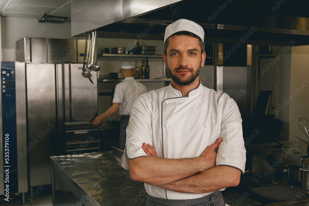Portrait of male chef in uniform standing on kitchen of restaurant with colleague on background Portrait of male chef in uniform standing on kitchen of restaurant with colleague on background