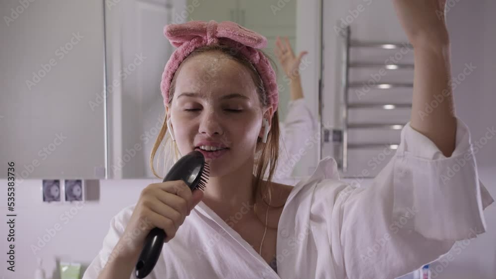 A young woman dances in the bathroom in wireless headphones and sings into a comb - looking in the camera