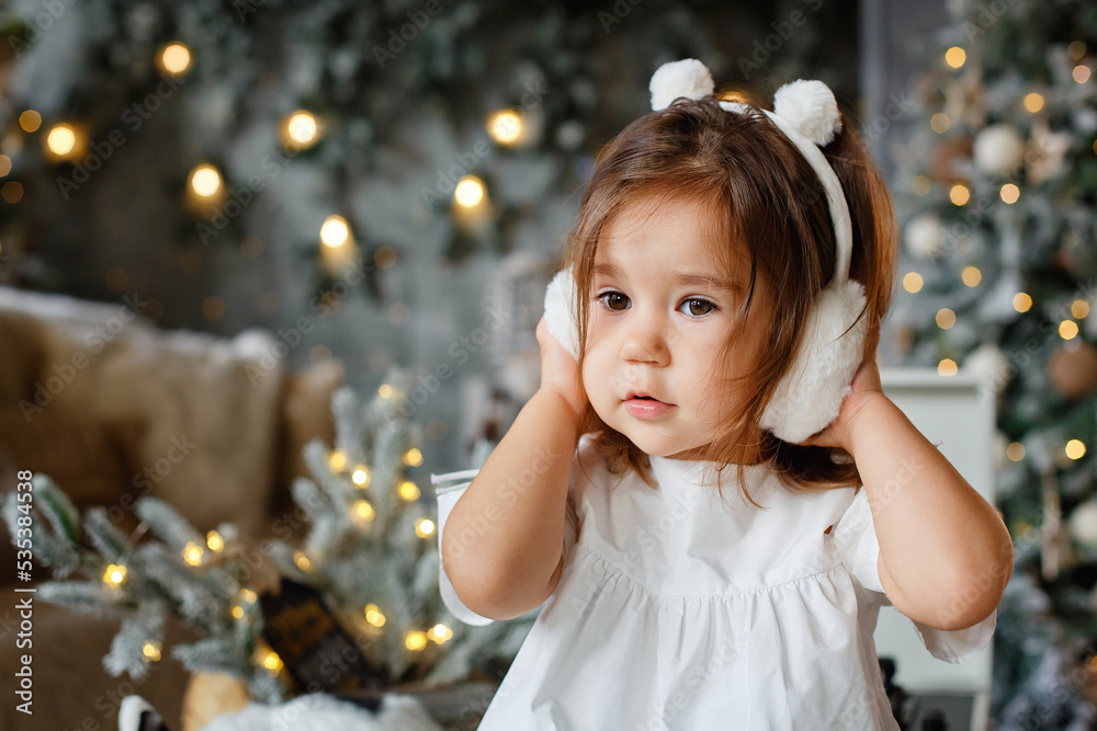 New Year's portrait of a girl, a cute child in white warm headphones on the background of a Christmas tree, Christmas mood, winter holiday, children's laughter. New Year's portrait of a girl, a cute child in white warm headphones on the background of a Christmas tree, Christmas mood, winter holiday, children's laughter.
