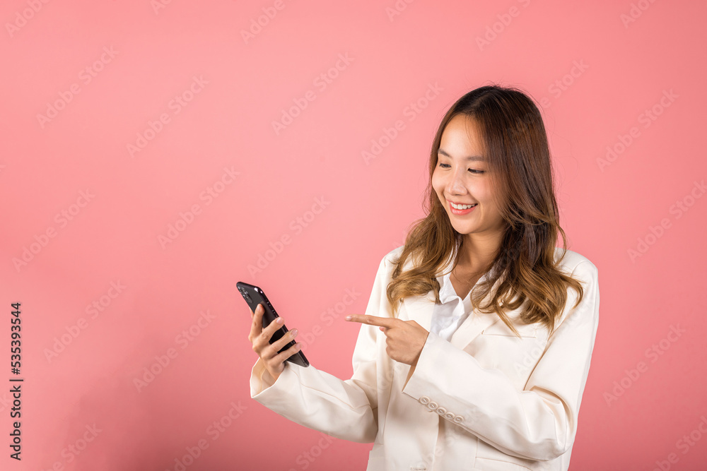 Portrait Asian happy beautiful young woman smile making finger pointing on screen smartphone in hand shopping online application looking to screen, studio shot isolated pink background with copy space Portrait Asian happy beautiful young woman smile making finger pointing on screen smartphone in hand shopping online application looking to screen, studio shot isolated pink background with copy space