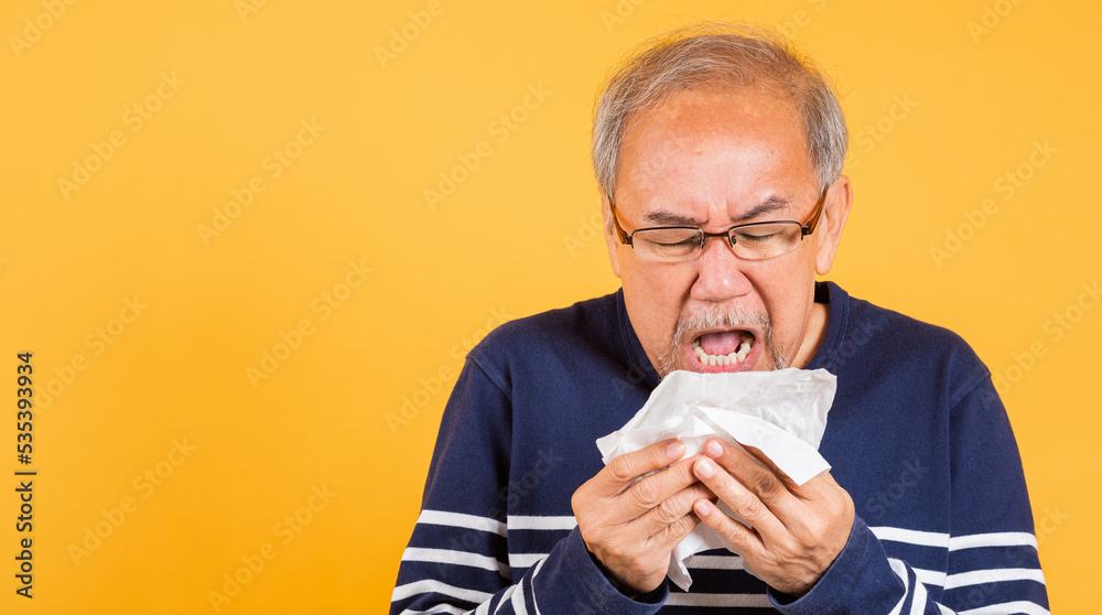 Asian elder man cold having flu and sneezing from sickness virus problem use tissue studio shot isolated on yellow background, Portrait senior old man blowing nose with tissue, pensioner unwell Asian elder man cold having flu and sneezing from sickness virus problem use tissue studio shot isolated on yellow background, Portrait senior old man blowing nose with tissue, pensioner unwell
