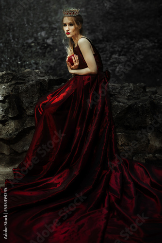 Woman in long red dress holding red apple sitting on rock tiara photo