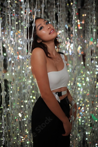 Woman in white spaghetti strap top and black skirt standing beside green christmas tree