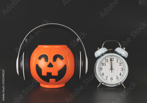 plastic halloween pumpkin  coverd with headphones and white vintage alarm clock 12pm., isolated on black  background with copy space. Halloween music or podcast. photo