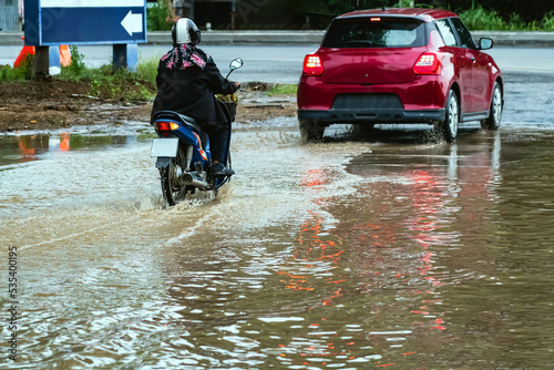 Woman ride motorcycle passing through flooded road. Riding motorbike on flooded road during flood caused by torrential rains. Flooded road with large puddle. Splash by motorcycle through flood water.
