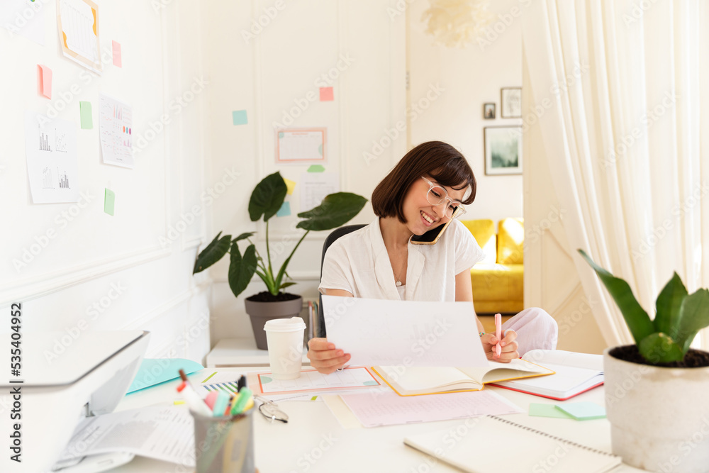 Happy young caucasian girl talking on phone with colleagues, making notes sitting at table. Brunette wears glasses, blouse at work. Office worker concept Happy young caucasian girl talking on phone with colleagues, making notes sitting at table. Brunette wears glasses, blouse at work. Office worker concept