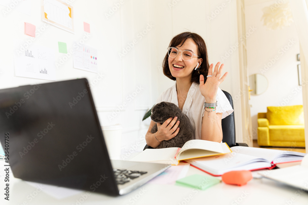 Pretty young caucasian girl is holding cat, waving her hand at laptop screen sitting at table. Brunette smiles, wears glasses and shirt. Concept use, leisure Pretty young caucasian girl is holding cat, waving her hand at laptop screen sitting at table. Brunette smiles, wears glasses and shirt. Concept use, leisure