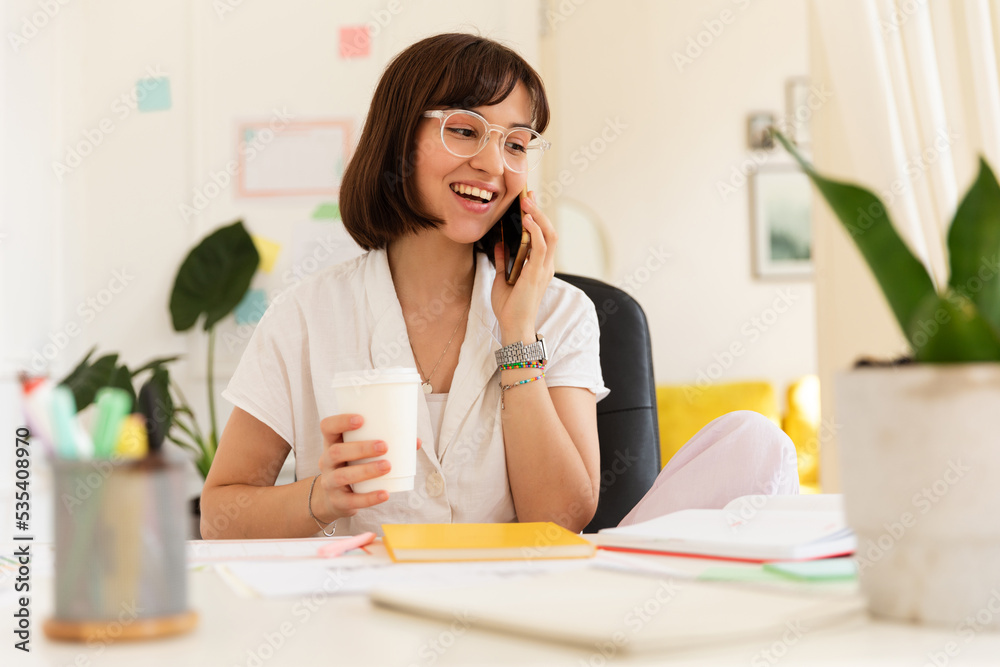 Joyful caucasian young woman talking on phone during coffee break sitting in office. Brunette with smile wears glasses and blouse. Concept of use Joyful caucasian young woman talking on phone during coffee break sitting in office. Brunette with smile wears glasses and blouse. Concept of use