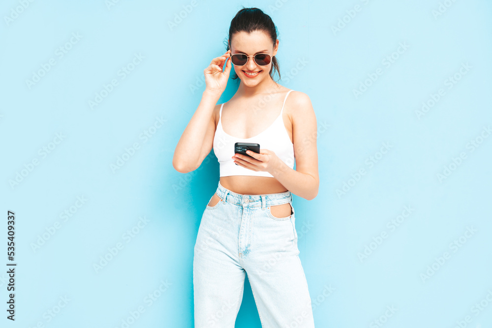 Young beautiful smiling female in trendy summer jeans. Sexy carefree woman posing near blue wall in studio. Positive brunette model looking at cellphone screen. Holding smartphone and using apps Young beautiful smiling female in trendy summer jeans. Sexy carefree woman posing near blue wall in studio. Positive brunette model looking at cellphone screen. Holding smartphone and using apps