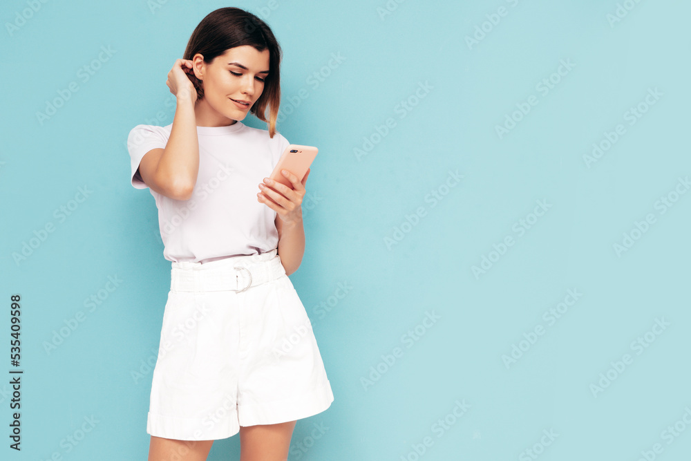 Young beautiful smiling female in trendy summer shorts clothes. Carefree woman posing near blue wall in studio. Sexy positive model Holding smartphone. Looking at cellphone screen. Using apps Young beautiful smiling female in trendy summer shorts clothes. Carefree woman posing near blue wall in studio. Sexy positive model Holding smartphone. Looking at cellphone screen. Using apps