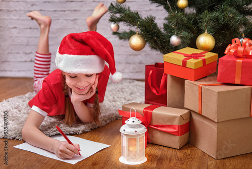 A happy smiling Christmas girl in a Christmas hat is writing a letter to Santa Claus, lying on the floor near the Christmas tree and Christmas gifts