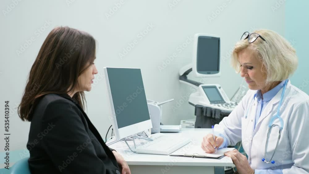 Mature blonde doctor writes down complaints in patient checklist form. Young woman describes complications to respiratory system after taking heavy antibiotics Mature blonde doctor writes down complaints in patient checklist form. Young woman describes complications to respiratory system after taking heavy antibiotics