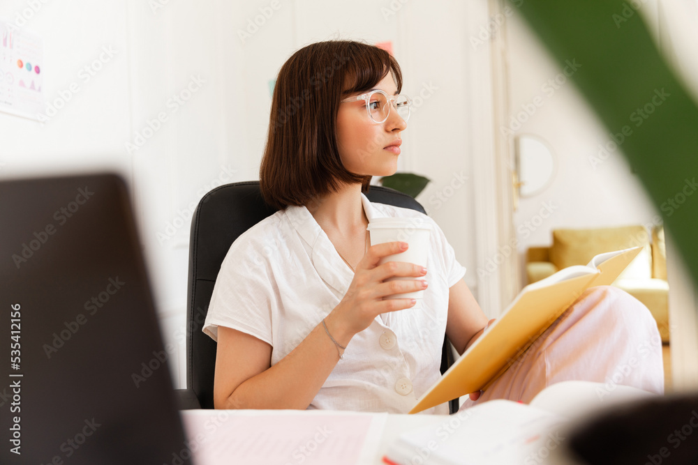 Calm caucasian young girl with notepad looks thoughtfully to side sitting indoors. Brunette wears glasses for vision and white blouse. Creativity and inspiration concept Calm caucasian young girl with notepad looks thoughtfully to side sitting indoors. Brunette wears glasses for vision and white blouse. Creativity and inspiration concept