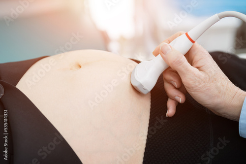 Doctor hand with ultrasonic device examines pregnant businesswoman. Ultrasound technician in white coat scans development and condition of embryo in abdomen, sunlight