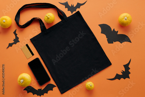 Top-down view shot of flat lay with black shopper bag, small pumpkins, smartphone, paper bats and bank card on orange surface photo