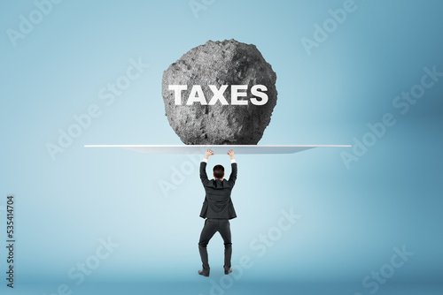 Burden of tax payments concept with businessman back view prevents huge stone from falling on his head with taxes sign on light blue background photo