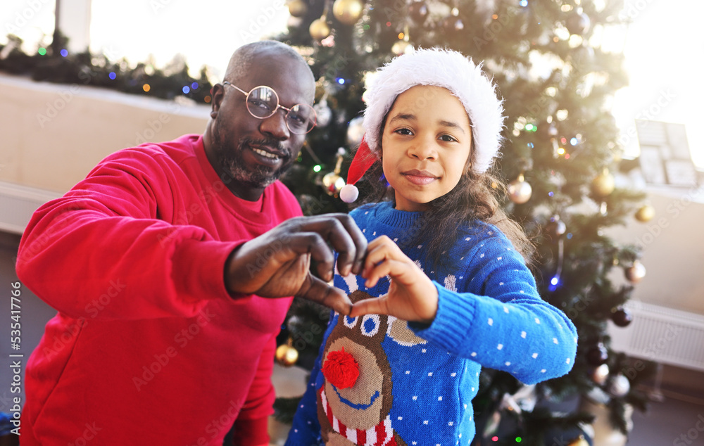dad and little African American daughter in New Year's costumes smile and make a heart out of their hands on the background of the Christmas tree.