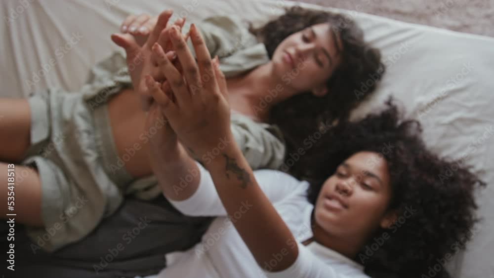 High angle slow motion selective focus of two young lesbian women in love relaxing on bed in morning enjoying touching each others hands High angle slow motion selective focus of two young lesbian women in love relaxing on bed in morning enjoying touching each others hands