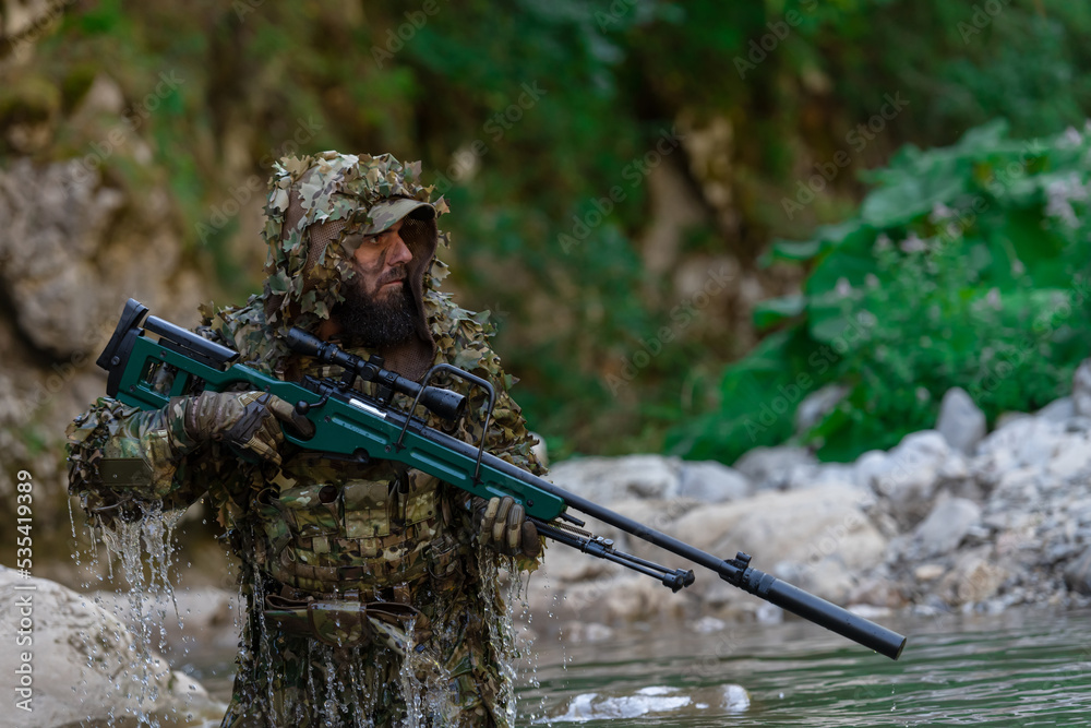 Naklejka premium A military man or airsoft player in a camouflage suit sneaking the river and aims from a sniper rifle to the side or to target.
