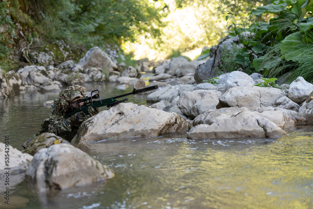 A military man or airsoft player in a camouflage suit sneaking the river and aims from a sniper rifle to the side or to target.