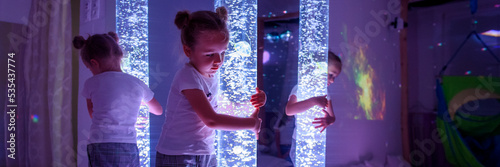 Child in therapy sensory stimulating room, snoezelen. Autistic child interacting with colored lights bubble tube lamp during therapy session. Web banner. photo