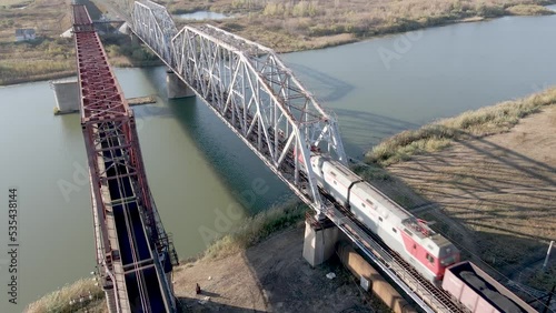An electric locomotive with freight cars with coal travels by rail on a railway bridge across the river. Transportation and delivery of goods in wagons.Transportation and logistics on the railway.